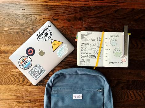 A laptop covered in fun stickers, a notebook full of information, and a backpack on a wooden table.