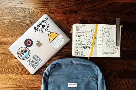 A laptop covered in fun stickers, a notebook full of information, and a backpack on a wooden table.