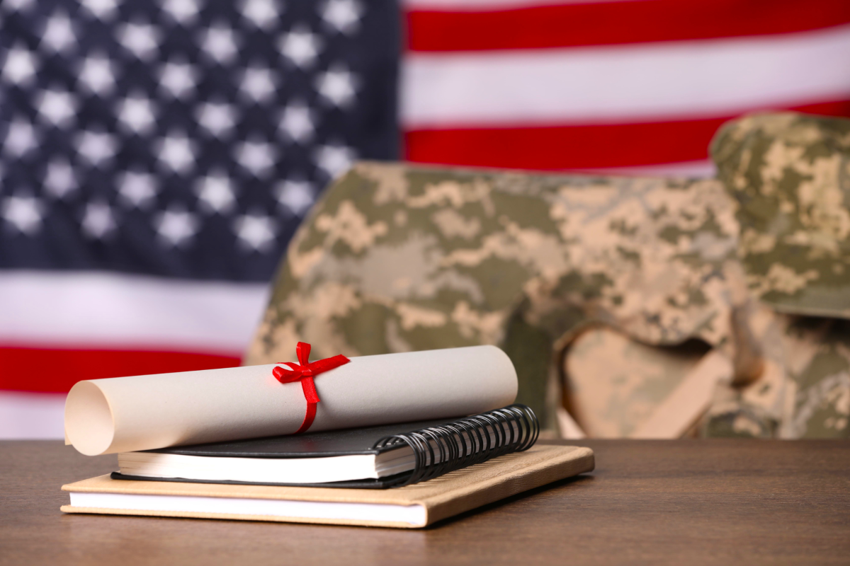 A diploma, notebook and textbook stacked on a desk. Behind the desk military fatigues are draped over a chair. An American Flag is displayed on the wall behind them.