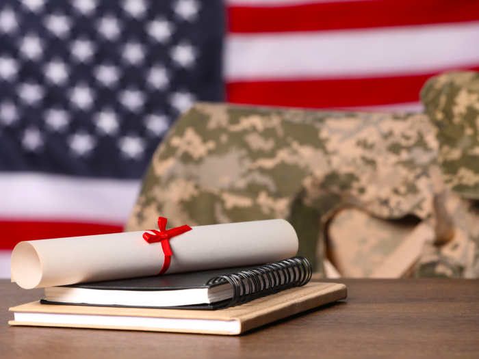 A diploma, notebook and textbook stacked on a desk. Behind the desk military fatigues are draped over a chair. An American Flag is displayed on the wall behind them.